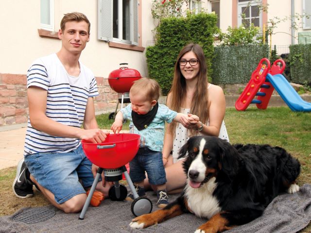 Familie mit kleinem Kind und Hund beim Picknick auf der Wiese vor dem Haus mit Grill