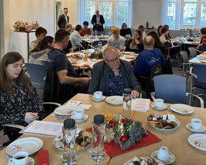 Menschen sitzen an gedeckten Tischen in einem hellen Veranstaltungsraum und nehmen gemeinsam an einer gemeinsamen Weihnachtsfeier mit Kaffee und Gebäck teil.